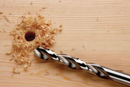 drill bit sitting on block of wood with a hole drilled in and saw dust surrounding the hole